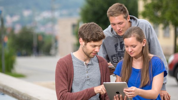 Drei Personen sitzen auf dem Rand eines Brunnens und schauen gemeinsam auf ein Tablet und füllen eine Online-Bewerbung für das Studium an der TU Ilmenau aus. Im Hintergrund sind verschwommen Gebäude der TU Ilmenau zu sehen.