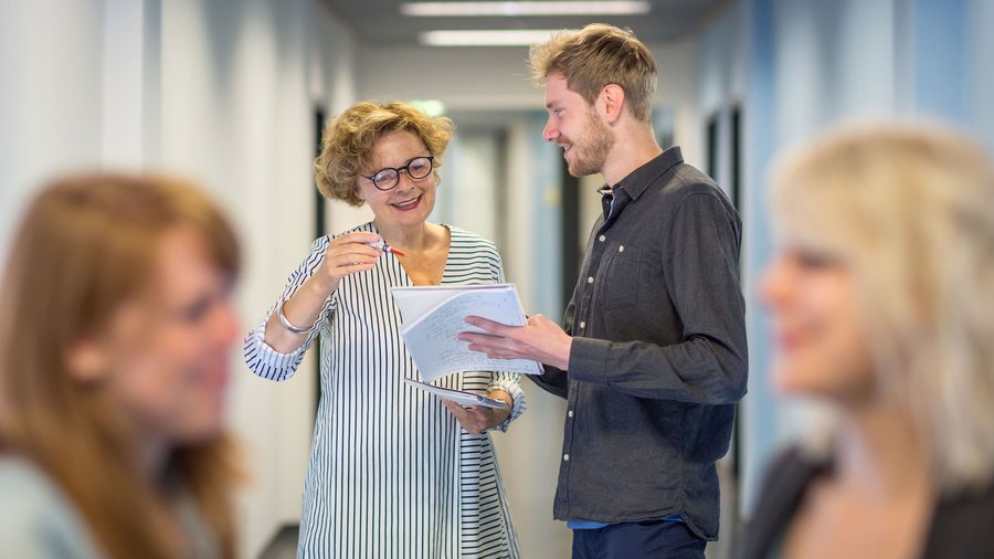 Professorin erläutert Studierendem Dokument, während sich im Vorfeld zwei Studierende unterhalten