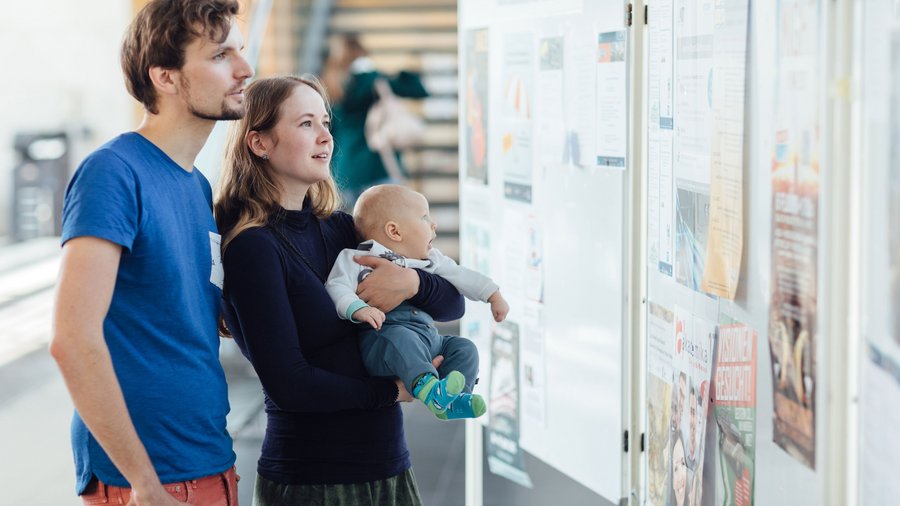 Mann und Frau mit Baby auf dem Arm schauen Aushänge an einer Pinnwand an.