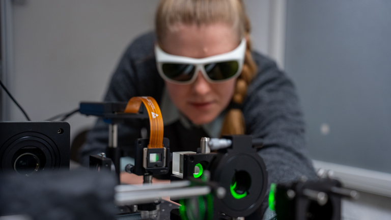 Wissenschaftlerin mit Schutzbrille hinter optischem Aufbau